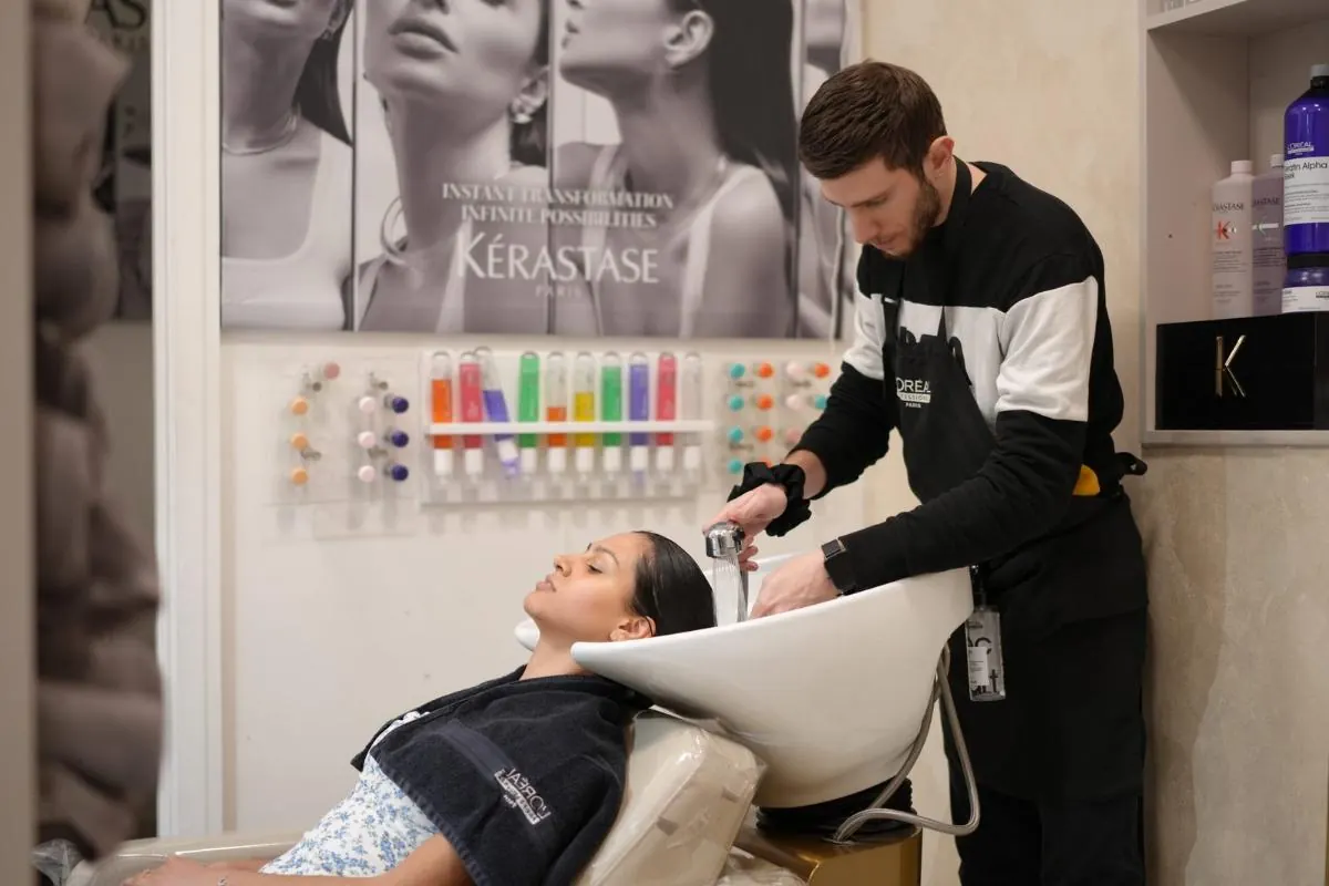 A stylist washing a client's hair at the basin, with Kérastase products on the wall